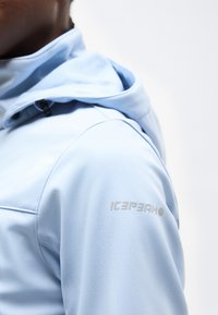 Άτομο που φοράει ανοιχτό μπλε μπουφάν softshell με κουκούλα, δείχνοντας το πάνω μέρος του βραχίονα και τον ώμο με το λογότυπο "ICEPEAK" στο μανίκι.