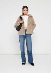 Veste beige douce avec une fermeture éclair à l'avant et une capuche, associée à un jean large bleu et un sac à bandoulière noir. Texture lisse et coupe décontractée.