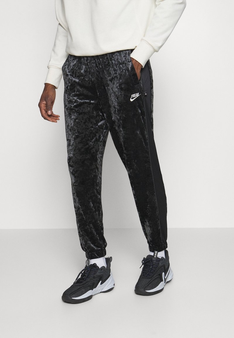 Pantalon de survêtement en velours noir avec poignets élastiques, panneaux latéraux en maille et un accent logo blanc. Porté avec des baskets noires et des chaussettes blanches.