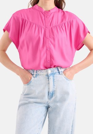 Femme portant un chemisier rose vif à manches courtes boutonné, rentré dans un jean taille haute bleu clair délavé, les mains dans les poches.