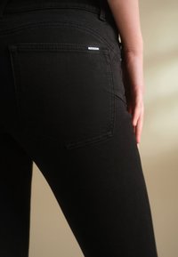 Gros plan d'une personne portant un jean noir avec une poche arrière visible et une petite étiquette métallique de marque, la main reposant à côté de la cuisse sur un fond neutre.