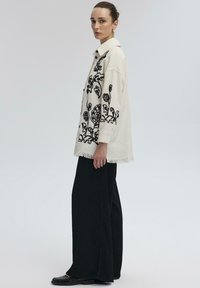 Crème oversized shirt met zwarte bloemenborduursels en een franjeszoom, gecombineerd met brede zwarte broeken en zwarte schoenen.