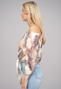 Blusa off-the-shoulder en crema con un patrón de hojas de palma marrones, que presenta un detalle de lazo en la cintura y mangas sueltas y abullonadas.