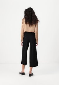 Blouse longue à manches longues en satin marron avec taille à volants, associée à un pantalon large noir. Portée avec des chaussures plates noires ornées d'accents circulaires.