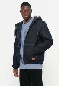 Denim jakna v temno modri barvi z kapuco, z dvema sprednjima žepoma, rebrastimi manšetami in rjavim usnjenim obročkom na robu. Nosi se čez modro pulover.