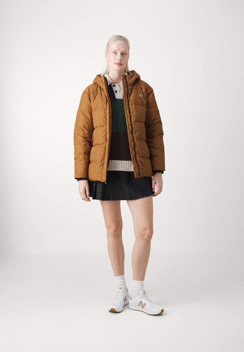 Doudoune marron avec une capuche, stylée sur un pull en tricot vert et marron, associée à une jupe noire et des baskets blanches.
