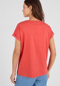 Camiseta de color rojo coral con mangas cortas enrolladas, un ajuste relajado y un dobladillo curvado. Presenta un inserto de encaje texturizado en las costuras laterales.