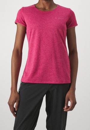 Henkilö, jolla on tiukka lyhythihainen pinkki urheilupaita ja väljät mustat housut, seisoo yksiväristä vaaleaa taustaa vasten.