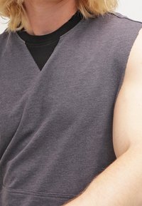 Top sin mangas de color gris oscuro con un diseño cruzado, hecho de suave tela de algodón. Presenta una capa interior negra contrastante en el cuello.