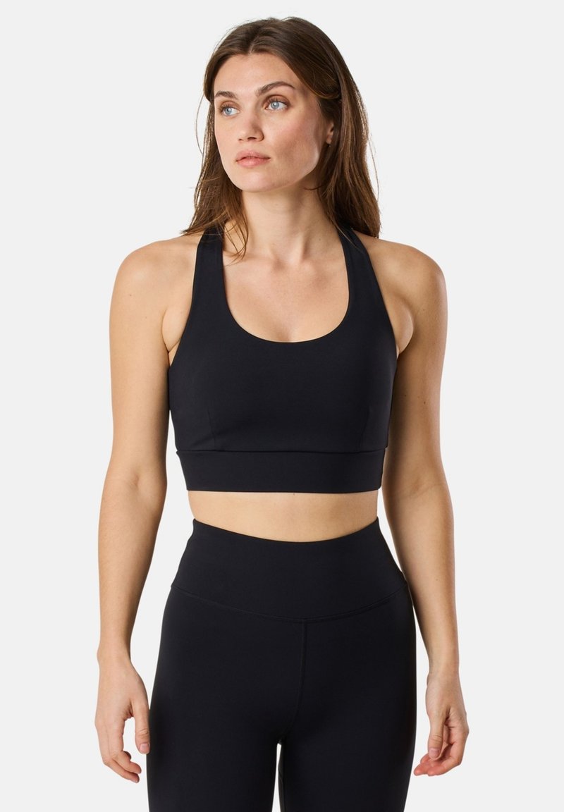 Kvinde iført sort sports-bh og leggings med høj talje, stående med armene afslappet, kiggende lidt til siden mod en ensfarvet baggrund.