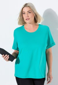 Žena s blond vlasy, která nosí tyrkysové tričko a drží černou láhev na vodu, stojí na bílém pozadí.