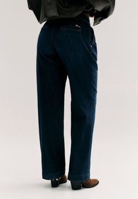 Personne portant un jean large bleu foncé, une veste en cuir noire et des chaussures marron à talons, debout devant un fond clair.