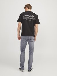 Schwarzes Baumwoll-T-Shirt mit weißem Text auf dem Rücken, der "Originals Studio Worldwide" liest, kombiniert mit hellgrauen Jeans und schwarzen Turnschuhen.
