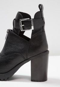 Czarne skórzane botki za kostkę z grubym obcasem, teksturowaną powierzchnią, srebrnym zapięciem oraz otwartymi bokami dla lepszej wentylacji.