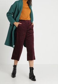 Cappotto lungo teal, dolcevita senape, pantaloni in corduroy bordeaux a gamba larga e stivaletti neri con tacco e lacci. Texture morbide e colori contrastanti.