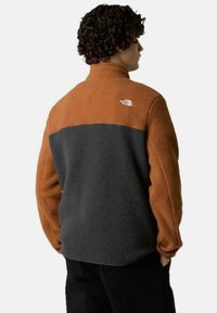 Chaqueta de forro polar con la parte superior marrón y la parte inferior gris oscuro. Tiene un cuello alto y un logo en la parte posterior. Textura suave.
