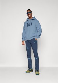 Felpa blu con logo, abbinata a pantaloni blu scuro con accenti azzurri. Il modello indossa occhiali da sole sportivi e scarpe pronte per il trail.