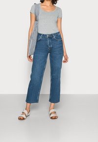 Šedé tričko s krátkým rukávem, modré džíny s vysokým pasem a rovnými nohavicemi, a bílé sandály. Součástí outfitu je černobílá pruhovaná taška přes rameno.