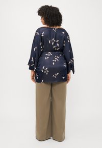 Blusa di seta blu navy con motivo floreale, maniche ampie e un fiocco in vita, abbinata a pantaloni beige chiaro a gamba larga.