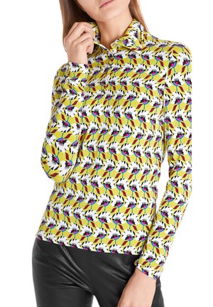 Femme portant un haut jaune à manches longues avec un motif floral blanc et noir audacieux et un pantalon en cuir noir.