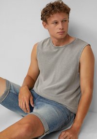 Top grigio senza maniche realizzato in tessuto morbido, abbinato a pantaloni in denim azzurri chiari con risvolti, seduto davanti a uno sfondo neutro.