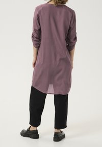 Tunique à manches longues, coupe décontractée en mauve, avec un col rond et des manches retroussées, assortie à un pantalon noir fuselé et des chaussures noires imposantes.