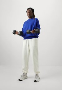 Niebieska bluza, białe spodnie dresowe, trzymająca czarne hantle. Szare buty sportowe o fakturowanej powierzchni. Prozy, codzienny strój do ćwiczeń.