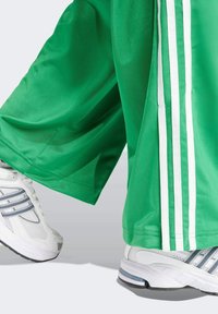 Groene sportbroek met witte strepen aan de zijkanten, gemaakt van glad synthetisch materiaal, gecombineerd met witte sportschoenen met grijze accenten.