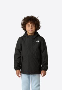 Veste noire imperméable avec capuche, poignets élastiques, deux poches latérales et éléments réfléchissants, portée avec un pantalon bleu clair.