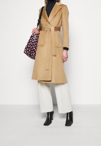 Frau trägt einen beigen Gürtelmantel, weiße weite Hosen, schwarze Stiefeletten, einen schwarzen Rollkragenpullover und hält eine gemusterte Schultertasche.