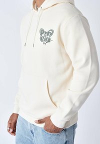 Sudadera de color crema hecha de tela suave, con un bolsillo frontal, capucha con cordón y un diseño gráfico en el pecho en gris.