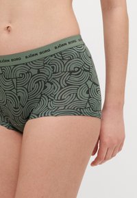 Shorts de boxeur en vert olive avec un motif en tourbillon abstrait noir, présentant une ceinture élastique marquée B.JÖRN BORG. Texture de tissu lisse.