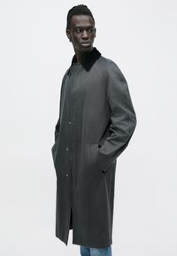 Grå, lang frakke med en glat struktur, trykknapper foran og sort krave. Har sidelommer og et afslappet fit.