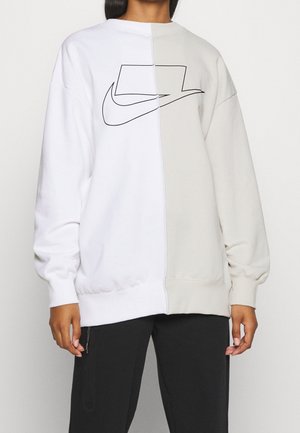 Personne portant un sweat-shirt moitié blanc, moitié beige clair avec un contour noir divisé du logo Nike swoosh et casquette sur la poitrine.