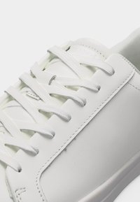 Gros plan sur une basket blanche montrant les coutures détaillées, les lacets texturés et la tige en cuir lisse sur un fond uni.