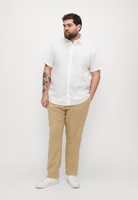 Cămășă albă cu mânecă scurtă și nasturi, pantaloni bej, pantofi albi, tatuaje vizibile pe braț, stând pe un fundal neutru.