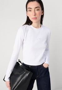 Donna in una maglietta bianca a maniche lunghe con il logo Calvin Klein, che tiene in mano una borsa nera, indossando jeans scuri, su uno sfondo neutro.