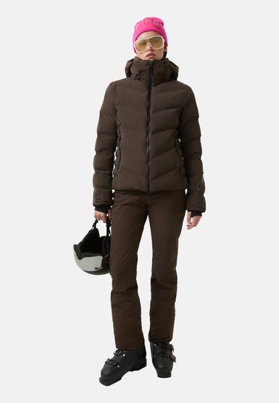 Braune Pufferjacke mit hohem Kragen, schwarzem Reißverschluss, braune Skihose, schwarze Skischuhe und eine rosa Mütze. Weißer Hintergrund, hält einen schwarzen Helm.