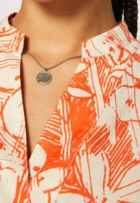 Gros plan d'une personne portant une chemise orange et blanche à motif et un collier en argent avec un pendentif circulaire.