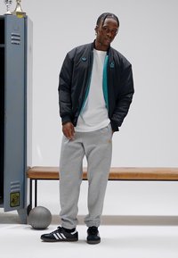 Μαύρο μπουφάν τύπου bomber με τυρκουάζ λεπτομέρειες, λευκό μπλουζάκι, γκρι φόρμα, και μαύρα αθλητικά παπούτσια με λευκές ρίγες· πόζα σε αθλητικό αποδυτήριο.