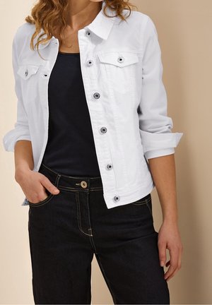 Kvinde iført en hvid denimjakke med knapper og opkørte ærmer over en sort skjorte og sorte jeans, stående foran en beige baggrund.