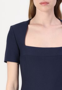 Temno modra obleka s kratkimi rokavi, kvadratnim izrezom, strukturiranim korzetom in gladko teksturo. Ima minimalno šivanje in čiste linije.