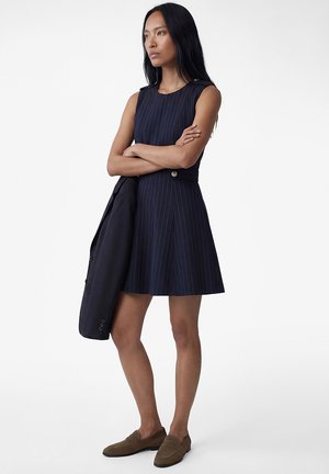 Mujer de pie con los brazos cruzados, vistiendo un vestido azul marino con rayas finas sin mangas, sosteniendo un blazer oscuro y con mocasines marrones sobre un fondo blanco.