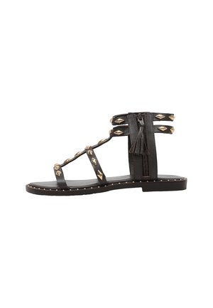 Sandalia plana negra con múltiples tiras adornadas con tachuelas doradas en forma de diamante y una cremallera lateral con tirador de borla.