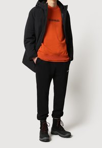 Chaqueta negra impermeable, sudadera naranja con cuello redondo y logo impreso, pantalones jogger negros y botas negras de cordones con suelas texturizadas.