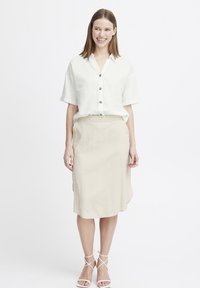 Camicia in lino bianca a maniche corte con bottoni, abbinata a una gonna beige di lunghezza al ginocchio, completata da sandali bianchi con cinturino.