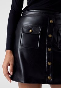 Czarna spódnica z czarnej skóry ekologicznej o dopasowanym kroju, z przodu zapinana na guziki oraz posiadająca dwie duże kieszenie z mosiężnymi zatrzaskami. Gładka faktura.