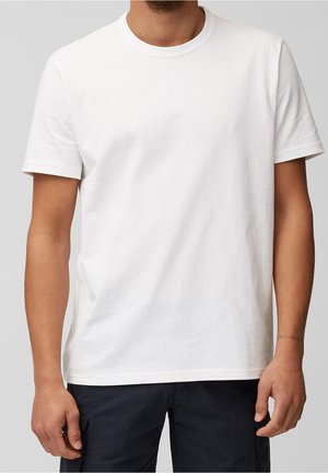 Mand iført en ensfarvet hvid t-shirt med korte ærmer og mørke bukser mod en neutral grå baggrund.