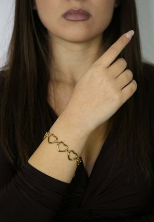 Goldenes Armband mit herzförmigen Gliedern am Handgelenk, mit glatter metallischer Textur und glänzendem Finish, getragen von einer Person, die mit dem Finger zeigt.