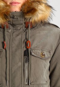 Personne portant une veste d'hiver grise avec capuche bordée de fourrure, cordons de serrage avec embouts couleur sable, une poche zippée et une poche à rabat à bouton.